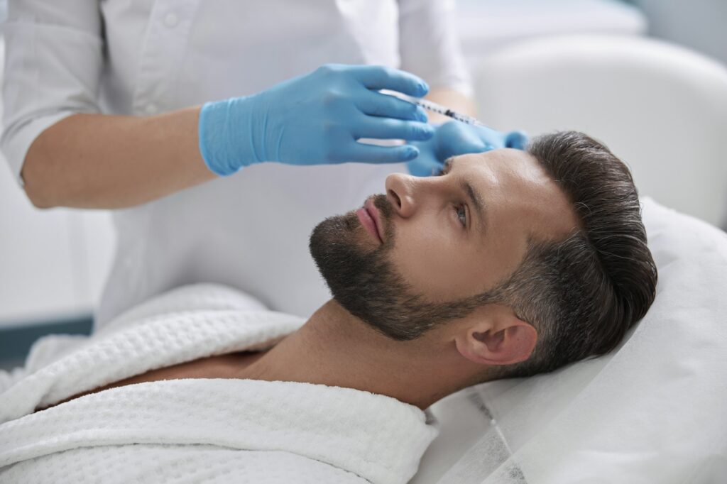 Lady cosmetologist holds syringe with filler above forehead of middle aged patient in salon