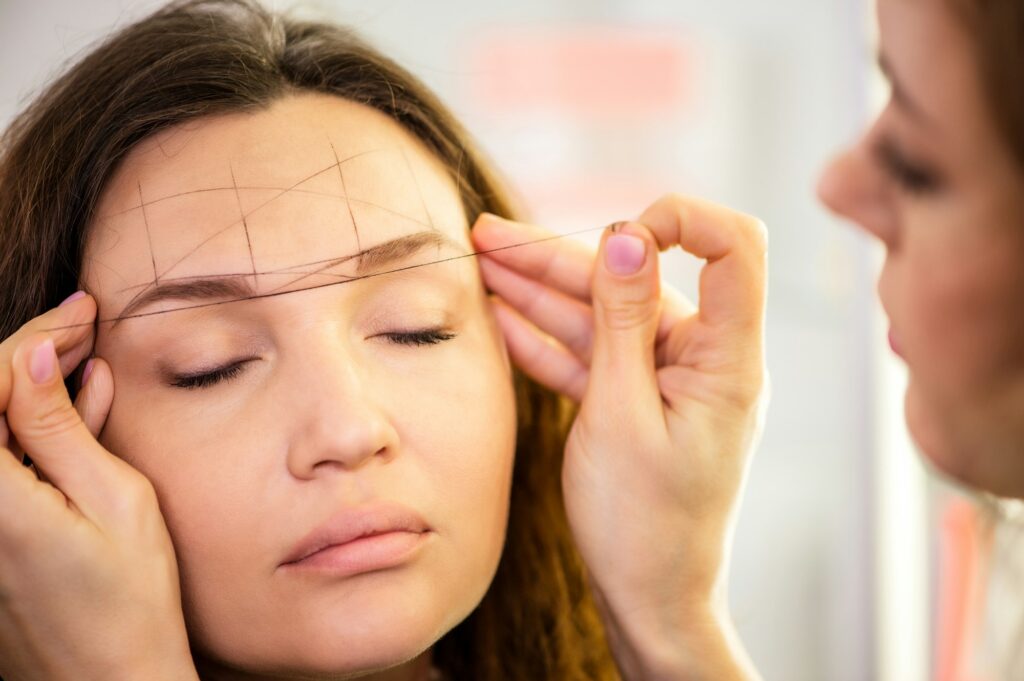 osmetologist forms the eyebrow line with a black thread on a female face.